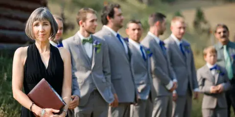 A woman in a black dress holding a book stands in front of a groom and groomsmen in grey suits at an outdoor wedding.