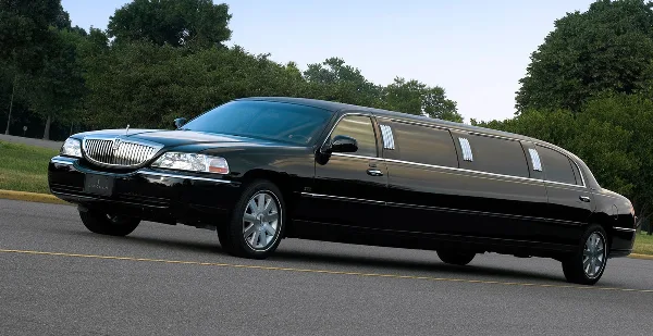 A black stretch limousine parked on a paved road with trees in the background.