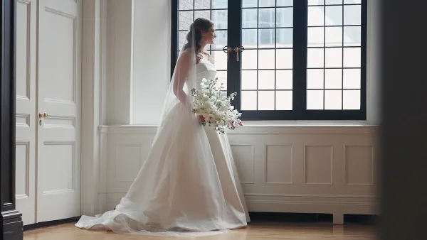 A bride in a white gown and veil stands by a large window holding a bouquet of white flowers.