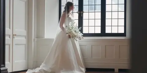 A bride in a white gown and veil stands by a large window holding a bouquet of white flowers.