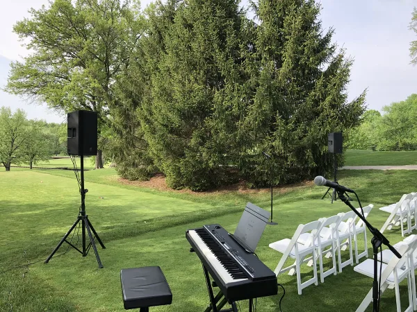 An outdoor wedding setup featuring a professional keyboard, microphone, and speakers arranged on a lawn next to rows of white chairs.