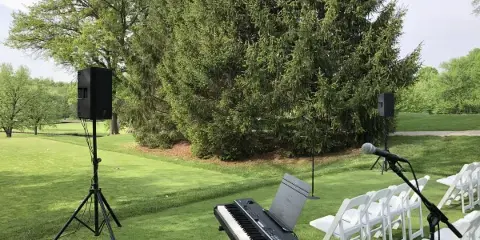 An outdoor wedding setup featuring a professional keyboard, microphone, and speakers arranged on a lawn next to rows of white chairs.