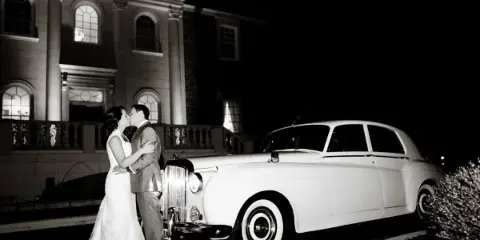 A black and white photograph of a bride and groom standing next to a white 1957 Bentley sedan in front of a large building.