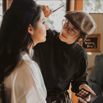 A professional makeup artist wearing a beret and glasses applies makeup to a client's face using a brush and a palette.
