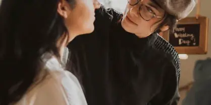 A professional makeup artist wearing a beret and glasses applies makeup to a client's face using a brush and a palette.