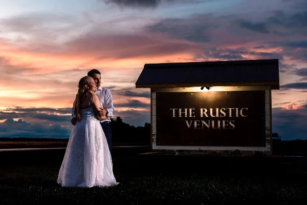 A bride and groom standing in front of the illuminated 'The Rustic Venues' sign at dusk.