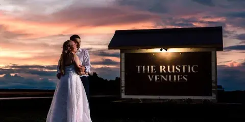 A bride and groom standing in front of the illuminated 'The Rustic Venues' sign at dusk.