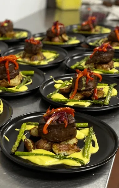 Multiple plated entrees with steak, potatoes, asparagus, and sauce arranged on a stainless steel counter.