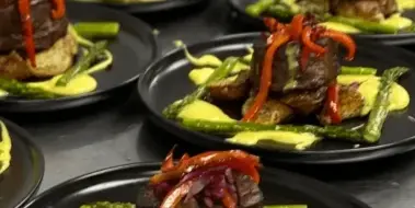 Multiple plated entrees with steak, potatoes, asparagus, and sauce arranged on a stainless steel counter.