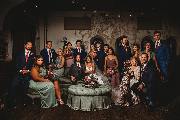 A formal group portrait of a wedding party, including the bride and groom, posing together in a rustic indoor venue.