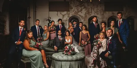 A formal group portrait of a wedding party, including the bride and groom, posing together in a rustic indoor venue.