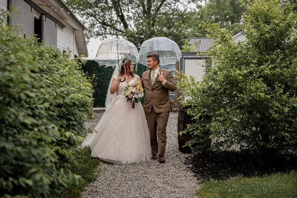 A bride and groom walking down a gravel path holding clear umbrellas, with the bride holding a bouquet.