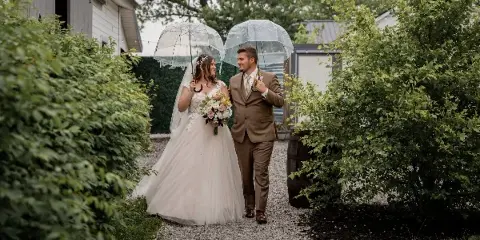 A bride and groom walking down a gravel path holding clear umbrellas, with the bride holding a bouquet.