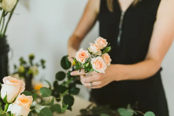 A florist arranging a bouquet of peach roses and greenery in a studio setting.