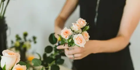 A florist arranging a bouquet of peach roses and greenery in a studio setting.