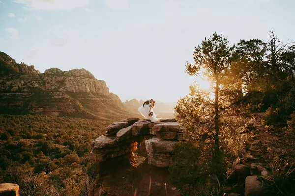 A bride and groom kissing on a rocky cliff overlooking a desert canyon at sunset.