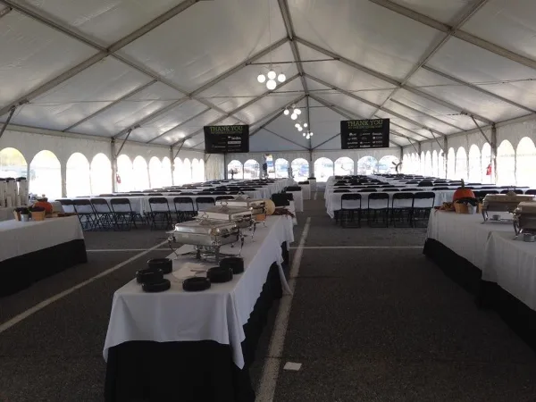 Interior of a large white event tent with rows of tables, chairs, linens, and buffet equipment set up for an event.