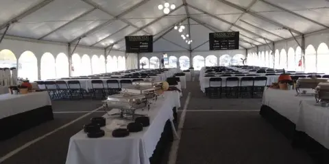 Interior of a large white event tent with rows of tables, chairs, linens, and buffet equipment set up for an event.