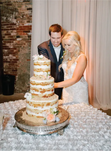 A bride and groom cutting a three-tiered semi-naked wedding cake at a reception.
