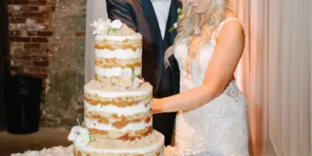 A bride and groom cutting a three-tiered semi-naked wedding cake at a reception.