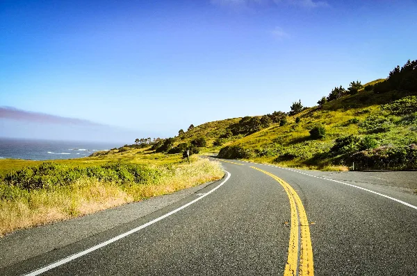 A winding paved road with yellow center lines curving through green coastal hills with the ocean visible in the distance.