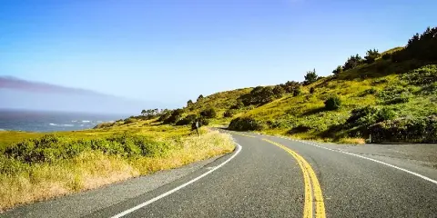 A winding paved road with yellow center lines curving through green coastal hills with the ocean visible in the distance.