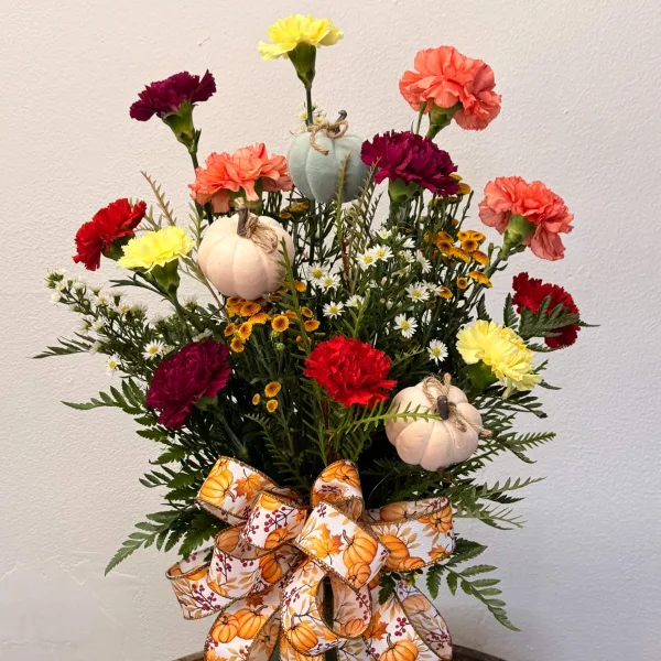 A fall-themed floral arrangement with red, yellow, and orange carnations, small white daisies, and decorative pumpkins tied with a ribbon.