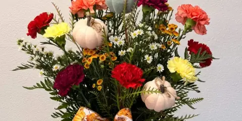 A fall-themed floral arrangement with red, yellow, and orange carnations, small white daisies, and decorative pumpkins tied with a ribbon.