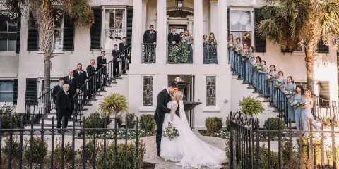 Wedding party posing on the exterior staircase and balcony of a historic white residence with columns.