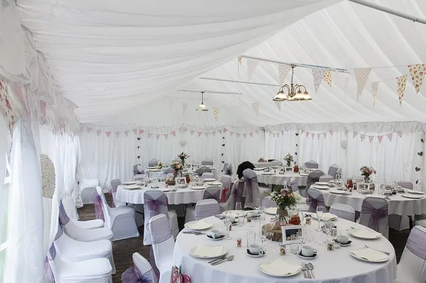 Interior of a white event tent featuring round tables with white linens, chairs with sashes, and decorative bunting.
