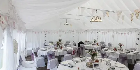 Interior of a white event tent featuring round tables with white linens, chairs with sashes, and decorative bunting.