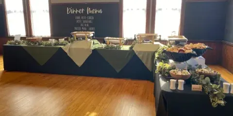 A catering buffet setup with a dinner menu board, food stations, and event decor in a venue.