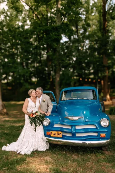 A bride and groom posing with a vintage blue pickup truck in a grassy outdoor area.