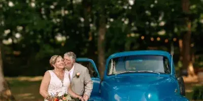 A bride and groom posing with a vintage blue pickup truck in a grassy outdoor area.