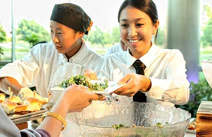 Two catering staff members in white uniforms serving a plate of food to a guest at an event.