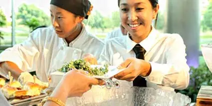 Two catering staff members in white uniforms serving a plate of food to a guest at an event.