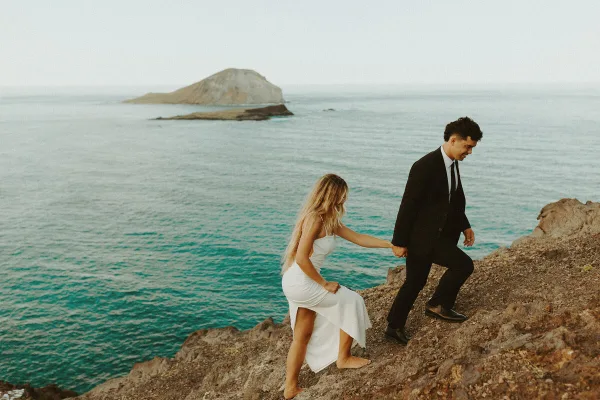 A bride and groom holding hands on a rocky coastal cliff overlooking the ocean and a distant island.