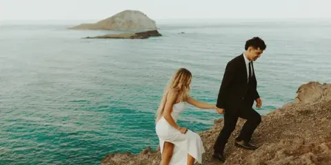 A bride and groom holding hands on a rocky coastal cliff overlooking the ocean and a distant island.