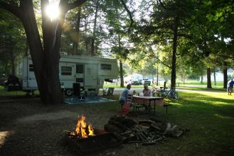 A couple sitting at a picnic table in a wooded campground with a camper and campfire.