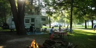 A couple sitting at a picnic table in a wooded campground with a camper and campfire.