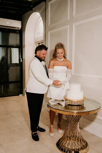 A bride and groom cutting a white wedding cake on a gold geometric pedestal table in a white-walled venue.