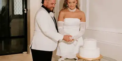 A bride and groom cutting a white wedding cake on a gold geometric pedestal table in a white-walled venue.