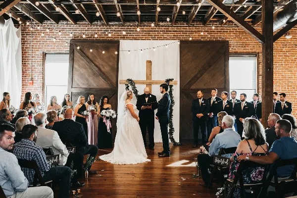 A wedding ceremony in progress at a renovated warehouse venue with exposed brick walls, wooden beams, and string lights.