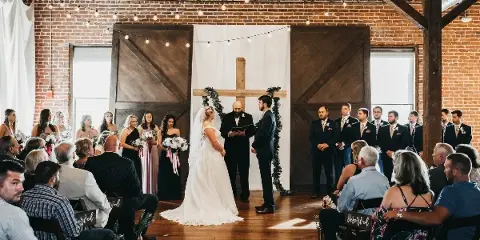 A wedding ceremony in progress at a renovated warehouse venue with exposed brick walls, wooden beams, and string lights.