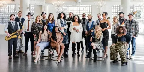 Group portrait of a large live band featuring multiple musicians with instruments including saxophones and guitars.