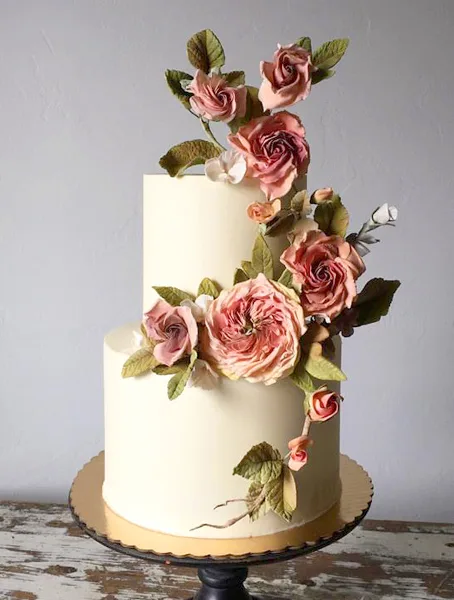 A two-tier white wedding cake decorated with cascading pink sugar roses and green leaves on a gold cake stand.