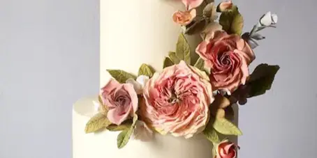 A two-tier white wedding cake decorated with cascading pink sugar roses and green leaves on a gold cake stand.