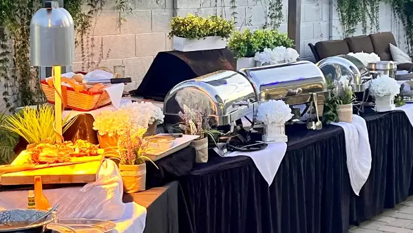 A full-service catering buffet setup featuring stainless steel chafing dishes, white floral arrangements, and black table linens.