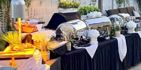 A full-service catering buffet setup featuring stainless steel chafing dishes, white floral arrangements, and black table linens.