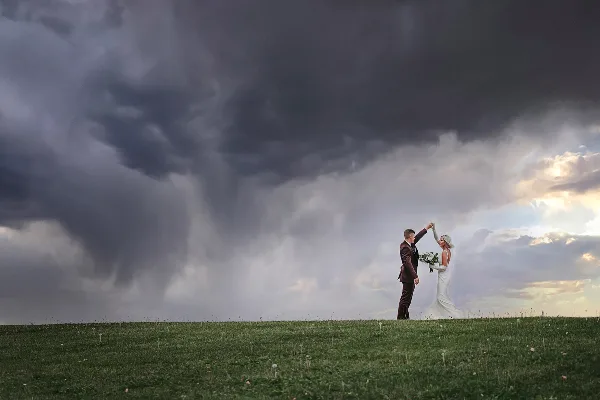A bride and groom dancing in a grassy field under a dramatic, cloudy sky.